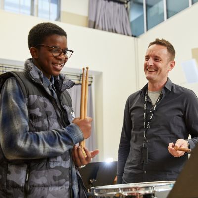A boy in a blue checked t-shirt and camouflaged gilet holds a pair of drum sticks. He is stood next to a man in a grey shirt. Both are smiling.